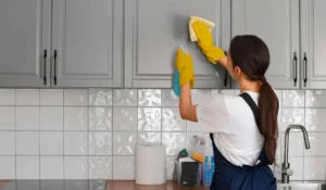 Professional residential cleaning services with a worker scrubbing gray cabinets in a modern kitchen setting.